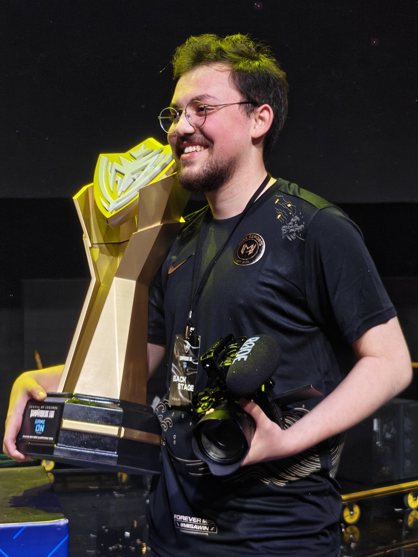 Ahmed Yıldız on an esports stage, holding a championship trophy and his cinema camera